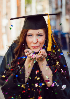 girl in graduation cap and gown blowing confetti from hands